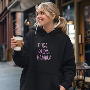 May include: Black hoodie with the text "Rosa sat, so Ruby could walk, so Kamala could run" in pink. The person in the image is holding a takeaway coffee cup.