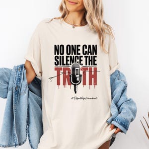 May include: A cream-coloured t-shirt featuring the text "NO ONE CAN SILENCE THE TRUTH" in black and red, with a microphone graphic. The hashtag "#Defendthefirstamendment" is printed at the bottom of the design.