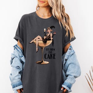 Puede incluir: Camiseta gris oscuro con una ilustración vintage de una mujer en una copa de champán, sosteniendo una copa de champán y un trozo de pastel. El texto "LET THEM EAT CAKE" está debajo. La camiseta tiene mangas enrolladas.