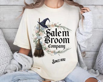 Könnte beinhalten: Ein weißes T-Shirt mit einem schwarzen grafischen Design aus einem Hexenhut, einem Besen und einem Blumengirlanden. Der Text lautet "Salem Broom Company Since 1692".