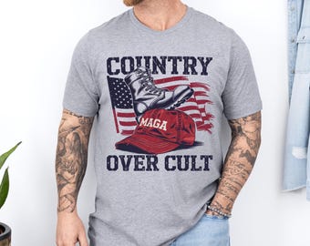 May include: A grey t-shirt featuring a graphic design of a military boot stepping on a red "MAGA" hat with an American flag in the background. The text "COUNTRY OVER CULT" is printed below the graphic.