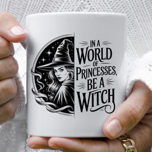 Może przedstawiać: Biały ceramiczny kubek z czarno-białą ilustracją wiedźmy i napisem "IN A WORLD OF PRINCESSES, BE A WITCH". Kubek jest trzymany przez osobę.