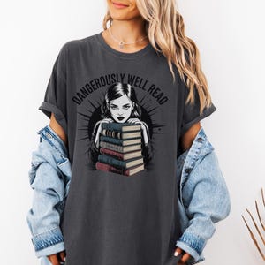 Könnte beinhalten: Dunkelgraues T-Shirt mit dem Text "DANGEROUSLY WELL READ" über einer Grafik einer Frau mit dunklem Haar, die ihre Arme auf einem Stapel Bücher abstützt. Die Bücher haben rote, blaue und beige Einbände.