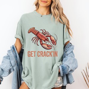 May include: A pale green t-shirt with a red and orange lobster design. The words "GET CRACK'N" are printed beneath the lobster. The shirt has short sleeves and a loose fit.