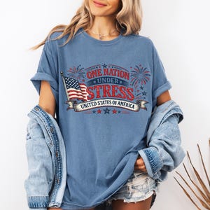 Puede incluir: Camiseta azul con el texto "ONE NATION UNDER STRESS UNITED STATES OF AMERICA" en rojo y azul. El diseño incluye una bandera americana y fuegos artificiales. La camiseta es de estilo casual.