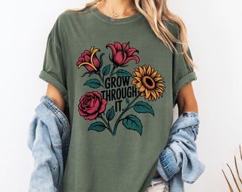 May include: An olive green t-shirt with a floral design including a rose and sunflower. The text "GROW THROUGH IT" is printed in black. The shirt has rolled-up sleeves and a loose fit. The shirt is made of a soft material.