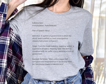 Puede incluir: Una camiseta gris de cuello redondo con la definición de "cacocracia" impresa en negro. El texto incluye la pronunciación, la parte de la oración, la definición, el origen y una frase de ejemplo. La camiseta se usa con jeans azules y una camisa a cuadros.