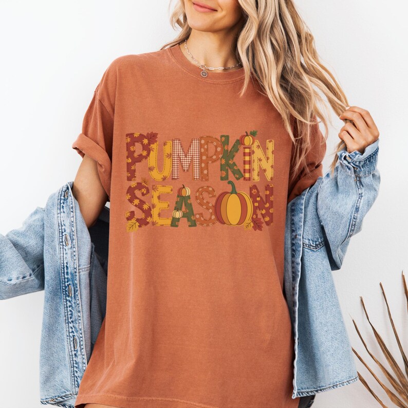 May include: Rust-colored t-shirt with the words "PUMPKIN SEASON" in a patchwork design. The letters are filled with fall-themed patterns and a pumpkin graphic. The shirt has short sleeves and a relaxed fit.