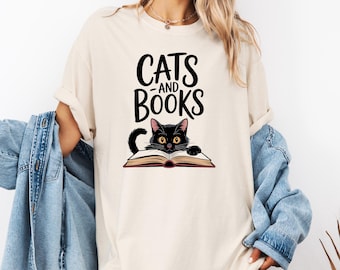 Könnte beinhalten: Cremefarbenes T-Shirt mit einer Illustration einer schwarzen Katze mit gelben Augen, die über ein offenes Buch blickt. Der Text "CATS and BOOKS" ist in fetter, schwarzer Schrift über der Katzenillustration gedruckt. Das T-Shirt hat hochgekrempelte Ärmel.