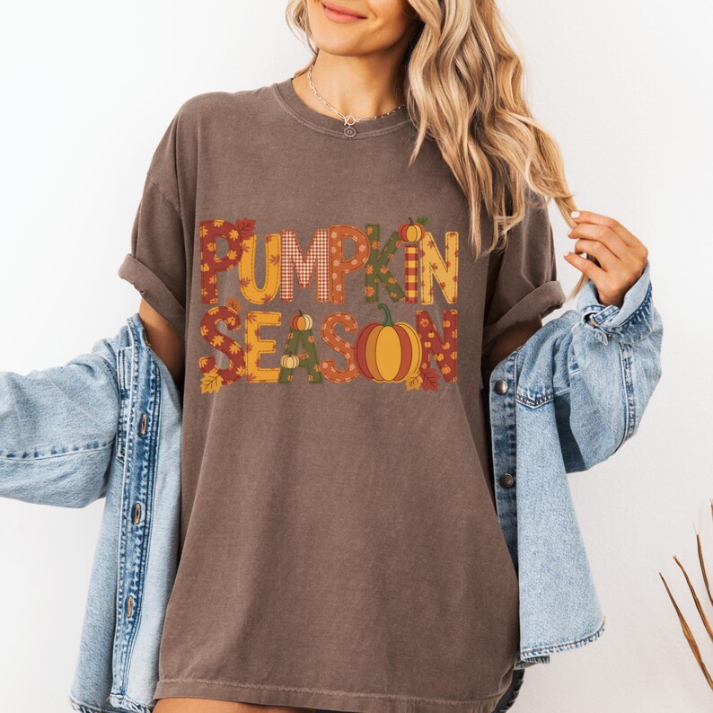 May include: A brown t-shirt with the words "PUMPKIN SEASON" in a fall-themed design. The letters are filled with patterns and a pumpkin graphic. The shirt is worn with a denim jacket.
