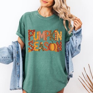 May include: A teal green t-shirt with the words "PUMPKIN SEASON" in a fall-themed design. The letters are filled with patterns and a pumpkin graphic. The shirt has short sleeves and a crew neck.