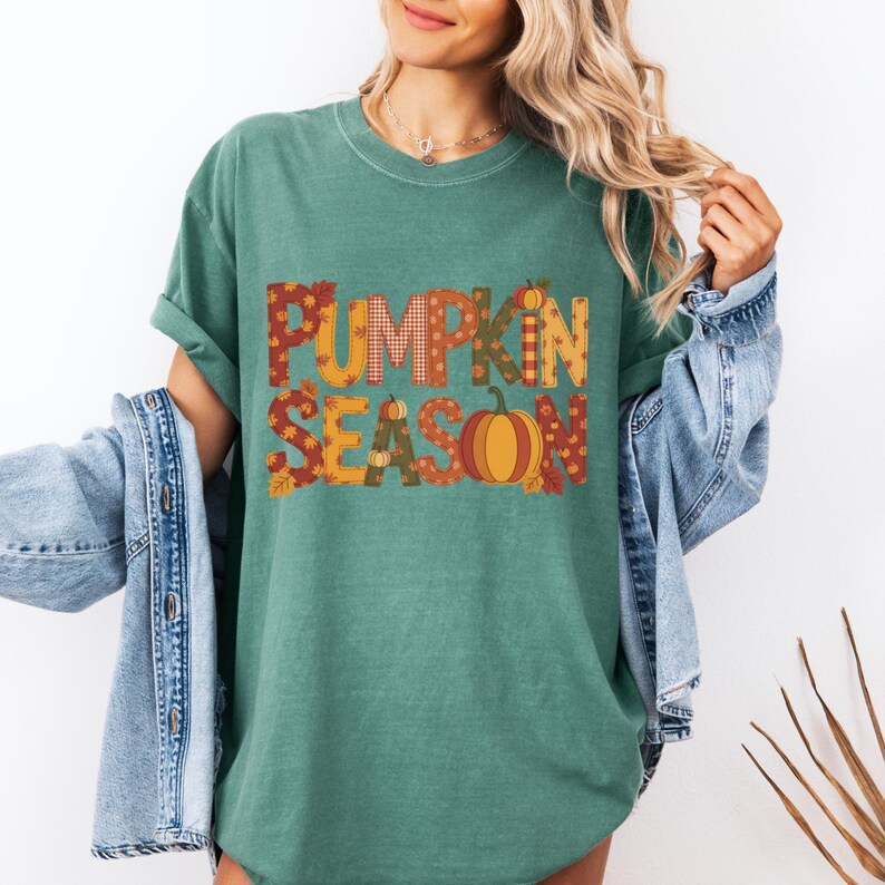 May include: A teal green t-shirt with the words "PUMPKIN SEASON" in a fall-themed design. The letters are filled with patterns and a pumpkin graphic. The shirt has short sleeves and a crew neck.