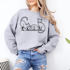 Op de afbeelding: Een lichtgrijs sweatshirt met een zwarte lijntekening van drie katten. Eén kat rekt zich uit, een andere slaapt opgerold en de derde zit rechtop. Het sweatshirt wordt door een persoon gedragen.