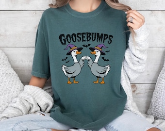 May include: Teal t-shirt with the word "GOOSEBUMPS" in a spooky font and a graphic of two geese wearing witch hats. Bats are scattered around the geese. The geese are grey with white feathers, orange feet, and black beaks.