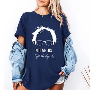 Könnte beinhalten: Marineblaues T-Shirt mit weißer Grafik eines Personensilhouette mit Brille und dem Text "NOT ME, US. Fight the oligarchy." Das T-Shirt hat einen lässigen Stil.