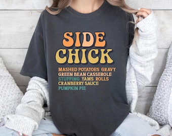 May include: Dark gray t-shirt with the text "SIDE CHICK" in orange and yellow lettering. Below the text are the words "MASHED POTATOES GRAVY GREEN BEAN CASSEROLE STUFFING YAMS ROLLS CRANBERRY SAUCE PUMPKIN PIE."