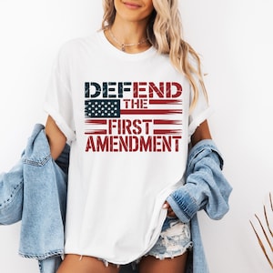 May include: White t-shirt featuring the text "DEFEND THE FIRST AMENDMENT" in a distressed font, with an American flag design. The shirt has short sleeves and a crew neck.