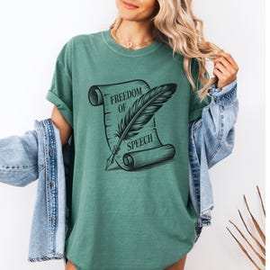 May include: A teal-colored t-shirt featuring a black graphic of a scroll with the words "FREEDOM OF SPEECH" and a quill pen. The shirt has short sleeves and a relaxed fit. A denim jacket is draped over the shoulders.