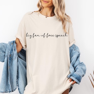 May include: Cream-coloured t-shirt with the phrase "big fan of free speech" in black script. The shirt has a relaxed fit and is paired with a blue denim jacket.