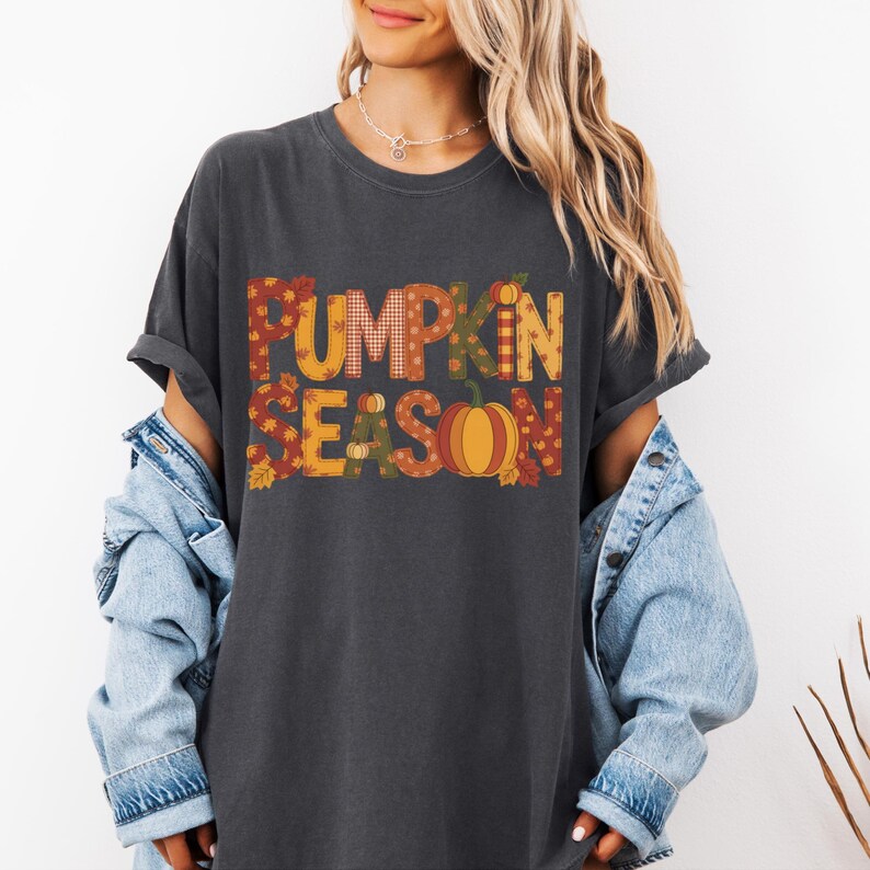 May include: Dark gray t-shirt with the words "PUMPKIN SEASON" in a fall-themed design. The letters are filled with plaid, leaves, and a pumpkin graphic. The shirt is worn with a denim jacket.