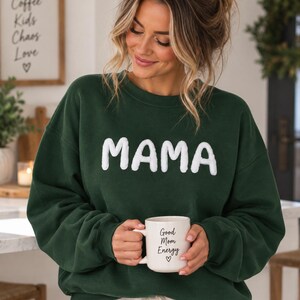 Può includere: Una felpa girocollo verde bosco scuro con la parola MAMA in grandi lettere bianche, testurizzate e dall'aspetto soffice sul petto. La felpa è abbinata a una tazza in ceramica bianca con la scritta Good Mom Energy e un cuore stampati sopra.