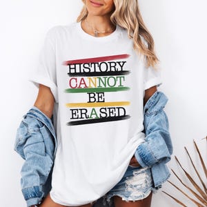 Può includere: T-shirt bianca con la scritta "HISTORY CANNOT BE ERASED" in nero, rosso, verde e giallo. Il testo è sovrapposto a tratti di pennello orizzontali. Un capo casual per tutti i giorni.