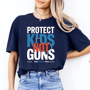 May include: Navy blue t-shirt with the text "PROTECT KIDS NOT GUNS" in white, blue, and red. The word "KIDS" features a blue Texas outline. The phrase "Gun Reform Now" is printed below the main text.