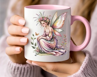 May include: White ceramic mug with a pink interior and handle. The mug features a colorful illustration of a fairy with floral accents, butterfly, and wings. The fairy is wearing a flowing gown. The mug is held by a person.
