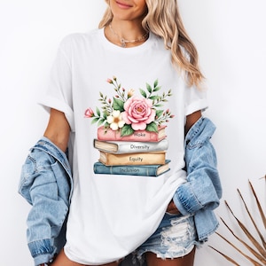 May include: White t-shirt featuring a stack of books and a pink flower illustration. The books have the words "Woke", "Diversity", "Equity", and "Inclusion" printed on them.