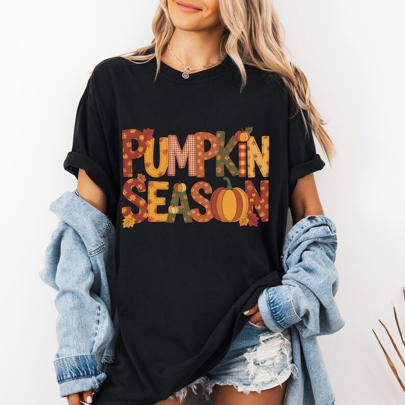 May include: Black t-shirt with the words "PUMPKIN SEASON" in a fall-themed design. The text features a pumpkin graphic and autumn leaf accents. The shirt is a casual, oversized fit.