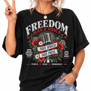 Puede incluir: Una camiseta negra con un gráfico de un micrófono vintage rodeado de amapolas rojas y margaritas blancas. El texto dice FREEDOM HAS A VOICE, FREE SPEECH, FREE PRESS, INFORM, EDUCATE, INVESTIGATE, CHALLENGE, INSPIRE, FACTS MATTER, TRUTH ENDURES, PEOPLE, IDEAS, DEMOCRACY.