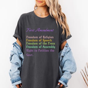 Puede incluir: Camiseta gris oscuro con el texto "First Amendment" en escritura púrpura. Debajo, la camiseta enumera las libertades de religión, expresión, prensa, reunión y el derecho a peticionar al gobierno en varios colores. Se usa con una chaqueta vaquera.
