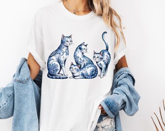 May include: White t-shirt with a graphic of four blue and white cats with floral patterns. The cats are arranged in a group, with one cat sitting upright and the others sitting or lying down.