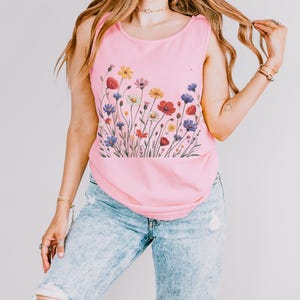 May include: A light pink tank top featuring a colorful floral design. The graphic includes red poppies, yellow daisies, purple and blue wildflowers. The tank top is paired with light blue denim jeans.