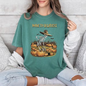 Könnte beinhalten: Türkisgrünes T-Shirt mit dem Text "BONE TO HARVEST" in goldfarbenen Buchstaben. Die Grafik zeigt ein Skelett mit Hut, das Kürbisse erntet. Ideal für Halloween oder den Herbst.