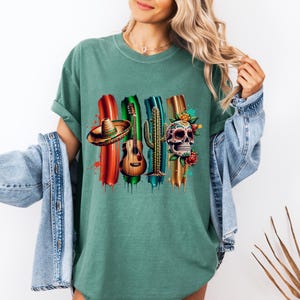 May include: Green t-shirt featuring a colorful graphic design with a sombrero, guitar, cactus, and decorated skull. The design includes red, green, blue, and gold brushstrokes, and floral accents. The shirt is a casual style.
