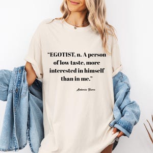 May include: Cream-colored t-shirt with the quote "EGOTIST, n. A person of low taste, more interested in himself than in me." by Ambrose Bierce in black text. The shirt is a casual style.