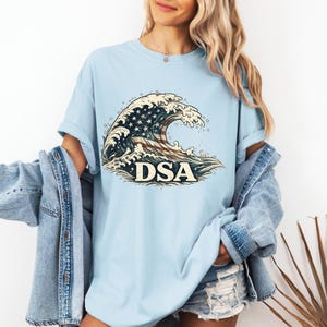 May include: A light blue t-shirt featuring a graphic of a large ocean wave patterned with the American flag and the letters DSA in a bold, serif font. The shirt has short sleeves and a crew neckline, worn with a denim jacket and denim shorts.