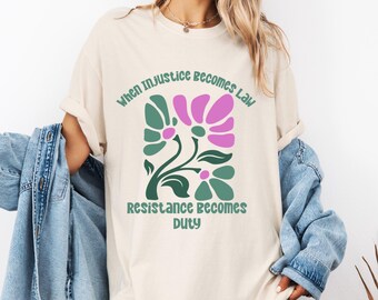 May include: Cream-colored t-shirt featuring a floral design in shades of green and pink, with the text "When Injustice Becomes Law, Resistance Becomes Duty" in green. The shirt has a relaxed fit.