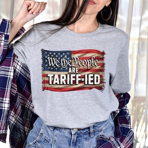 May include: A gray t-shirt with a distressed American flag design and the text "We the People are Tariff-ied".