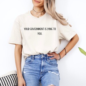 Puede incluir: Una camiseta color crema con el texto "YOUR GOVERNMENT IS LYING TO YOU" en negro. La camiseta se combina con vaqueros azules. La imagen está tomada con luz natural.