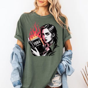 Könnte beinhalten: Olivgrünes T-Shirt mit einer Grafik einer Frau, die ein Buch mit dem Titel "1984" von Orwell hält, mit Flammen. Die Frau hat einen ernsten Gesichtsausdruck. Der Text und die Grafik sind in Schwarz und Weiß.