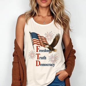 May include: A cream-coloured sleeveless tank top featuring a graphic print of an American flag, a bald eagle in flight, and red and blue fireworks. The text "Freedom Truth Democracy" is printed vertically in red and blue lettering on the front.