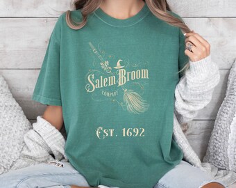 Könnte beinhalten: Ein teal-grünes T-Shirt mit dem Text "Salem Broom Company" und "EST. 1692" im Vintage-Stil. Das Design umfasst einen Hexenhut, einen Besen und andere dekorative Elemente. Das Shirt hat eine lockere Passform.