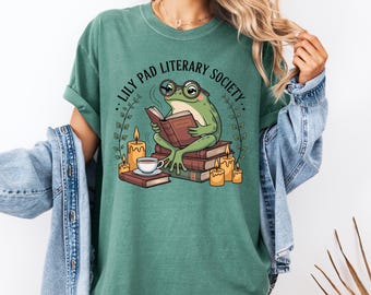 Może przedstawiać: Zielona koszulka z grafiką żaby w okularach czytającej książkę, otoczonej świecami i książkami. Tekst "LILY PAD LITERARY SOCIETY" jest wydrukowany nad żabą. Koszulka jest w jednolitym zielonym kolorze.