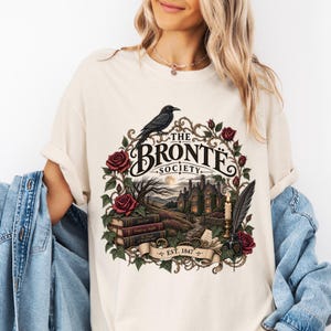 Puede incluir: Una camiseta color crema con un estampado gráfico de un cuervo posado sobre el texto The Bronte Society Est. 1847. El diseño incluye una mansión gótica, una pila de libros, rosas rojas, una vela encendida y una pluma en un tintero.