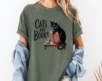 Könnte beinhalten: Olivgrünes T-Shirt mit dem Text "Cats and Books" und einer Grafik einer schwarzen Katze, die ein Buch liest. Ein kleiner Tisch trägt das Buch und eine Tasse Kaffee. Die Katze hat grüne Augen.
