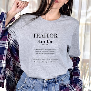 May include: A grey t-shirt featuring the word "TRAITOR" in black, with its phonetic pronunciation and definition. The definition states: "A person who betrays a friend, country, principle, or trust. one who commits treason."