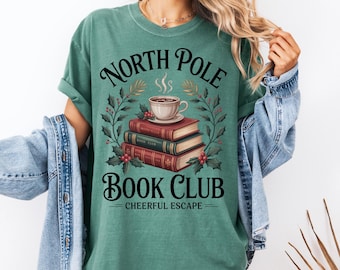 Könnte beinhalten: Ein teal-grünes T-Shirt mit dem Schriftzug "North Pole Book Club" in einer dekorativen Schriftart. Das Design umfasst einen Stapel Bücher, eine Tasse Kaffee und Stechpalmenblätter. Der Satz "Cheerful Escape" ist darunter gedruckt.