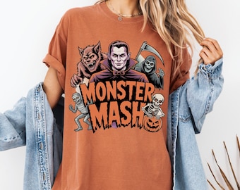 May include: Rust-coloured t-shirt with a graphic of Halloween monsters: a werewolf, Dracula, the Grim Reaper, a mummy, and a skeleton. The words "Monster Mash" are printed in orange and black.